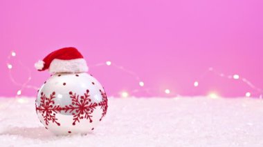 Kar ve yanıp sönen ışıklar ile birlikte Pembe Arkaplanda Noel Baba Şapkası olan bir Beyaz ve Kırmızı Noel Süslemesi