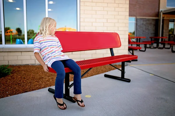 一个寂寞的小女孩正坐在学校的长椅上等着一个朋友和她一起在操场上玩耍 — 图库照片