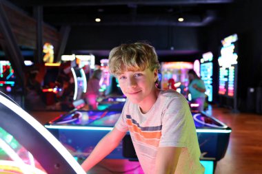 A 14 year old teenage boy is playing a game at a video arcade with neon lights. clipart