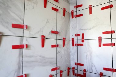 A wall of tile work is being done on a shower and red leveling spacers are dividing the tiles. clipart