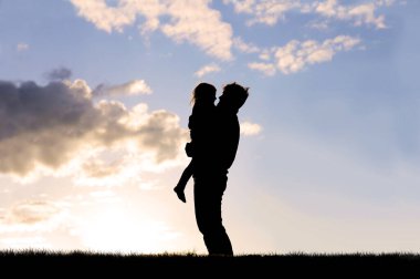 Mutlu, genç bir babanın silueti küçük kızını kucağına alıyor ve ona sarılıyor..