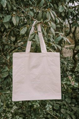 A blank canvas tote bag hanging from a tree branch in a lush green setting. clipart