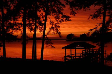 Silhouette of a dock at sunset with vibrant orange sky and tranquil water. clipart