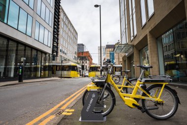 Yellow bicycles lined up on a street in Manchester for hire seen next to double yellow lines in February 2023 as two trams pass is the background.