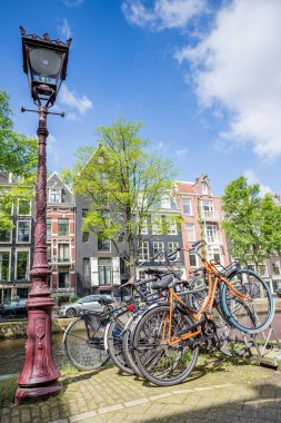 Hollanda, Amsterdam 'da Reguliersgracht kanalı boyunca park etmiş kırmızı bir sokak lambasının yanına park etmiş renkli bisikletler 28 Mayıs 2024' te ince kanal evleri tarafından desteklenmektedir..