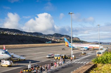 İngiliz turistler 12 Ağustos 2024 'te Madeira' daki Funchal havaalanındaki eşikten geçerek eve dönüş uçağını yakalarken bir Alman Akbaba uçağı arka planda kalkıyor..