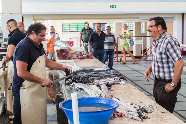 Stock image Two men talk over a counter as the fishmonger uses his knife to prepare a Black Scabbard fish in Mercado dos Lavradores market for sale.  Pictured on 6 August 2024 in the centre of Funchal, Madeira.