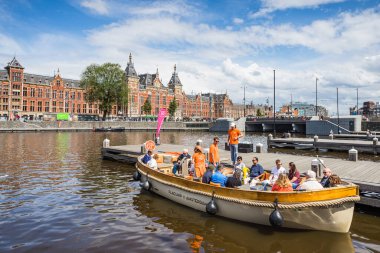 27 Mayıs 2024 'te Hollanda' da Amsterdam Centraal 'ın önünde bir turist teknesi, şehir çevresinde bir kanal gezisine çıkmak için turistlerle dolup taşar..