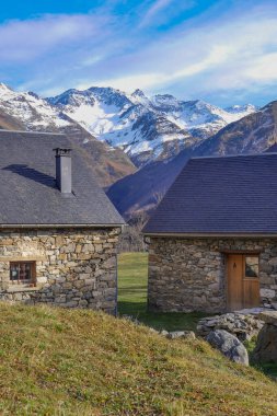 İki büyüleyici taş ev, parlak mavi bir gökyüzünün altında, yüksek karla kaplı dağlarla çevrili, sakin bir vadide yuva yapar. Resimli ortam sükunet yayıyor..
