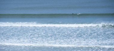 İki sörfçü, sakin bir günde Biscarrosse Sahili 'nde dalgalanan dalgalarda dolaşır. Sakin sular masmavi gökyüzünü yansıtıyor ve mükemmel bir sörf ortamı yaratıyor..