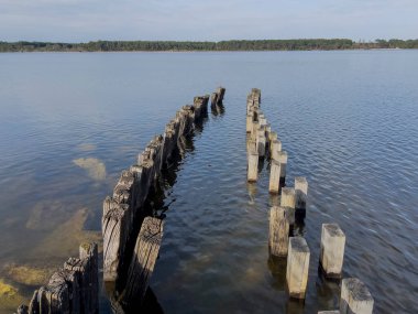 Yıpranmış ahşap sütunlar, bereketli yeşillik ve parlak mavi gökyüzü çerçeveli sakin gölün sularında çarpıcı bir desen oluşturarak ziyaretçileri doğal güzelliği keşfetmeye davet ediyor..