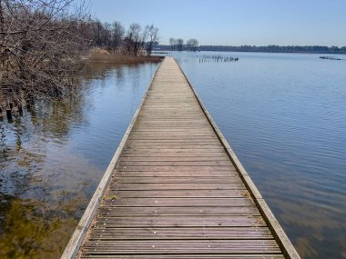 Biscarosse gölünün sakin suları üzerinde uzun tahta bir yürüyüş yolu uzanır. Çevresindeki bitki örtüsü ve uzun sazlıklar berrak mavi gökyüzünün altında barışçıl bir atmosfer yaratıyor..