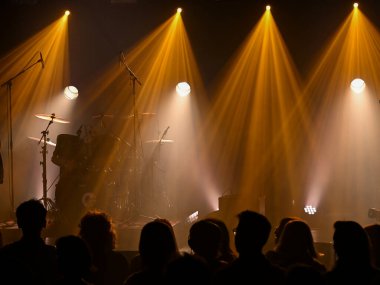 Müzisyenler enstrümanlarını ayarlarken parlak ışıklar sahneyi aydınlatıyor. Kalabalık enerji ve heyecanla dolu canlı bir konser deneyimini heyecanla bekliyor..