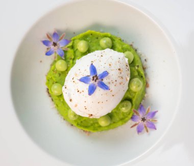 Taze bezelye püresi güzel bir şekilde kaplanmış, ortasında haşlanmış bir yumurta ve borage çiçekleriyle süslenmiş. Yemek canlı renkler ve dokular sergiliyor, her bahar menüsü için mükemmel..