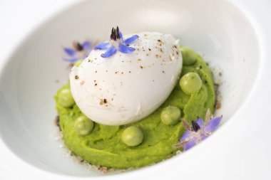Taze bezelye püresi güzel bir şekilde kaplanmış, ortasında haşlanmış bir yumurta ve borage çiçekleriyle süslenmiş. Yemek canlı renkler ve dokular sergiliyor, her bahar menüsü için mükemmel..