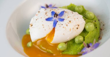 Taze bezelye püresi güzel bir şekilde kaplanmış, ortasında haşlanmış bir yumurta ve borage çiçekleriyle süslenmiş. Yemek canlı renkler ve dokular sergiliyor, her bahar menüsü için mükemmel..