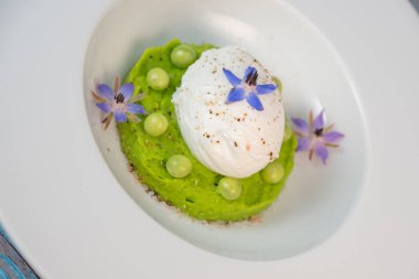 Taze bezelye püresi güzel bir şekilde kaplanmış, ortasında haşlanmış bir yumurta ve borage çiçekleriyle süslenmiş. Yemek canlı renkler ve dokular sergiliyor, her bahar menüsü için mükemmel..