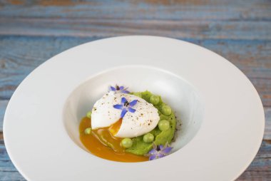 Taze bezelye püresi güzel bir şekilde kaplanmış, ortasında haşlanmış bir yumurta ve borage çiçekleriyle süslenmiş. Yemek canlı renkler ve dokular sergiliyor, her bahar menüsü için mükemmel..