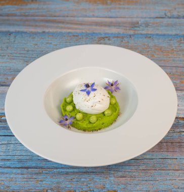 Taze bezelye püresi güzel bir şekilde kaplanmış, ortasında haşlanmış bir yumurta ve borage çiçekleriyle süslenmiş. Yemek canlı renkler ve dokular sergiliyor, her bahar menüsü için mükemmel..