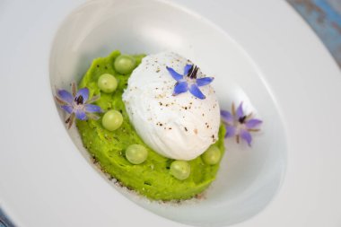 Taze bezelye püresi güzel bir şekilde kaplanmış, ortasında haşlanmış bir yumurta ve borage çiçekleriyle süslenmiş. Yemek canlı renkler ve dokular sergiliyor, her bahar menüsü için mükemmel..