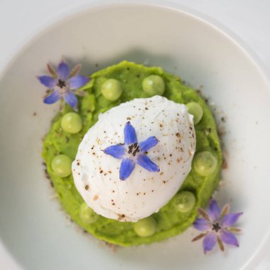 Taze bezelye püresi güzel bir şekilde kaplanmış, ortasında haşlanmış bir yumurta ve borage çiçekleriyle süslenmiş. Yemek canlı renkler ve dokular sergiliyor, her bahar menüsü için mükemmel..
