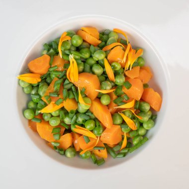 Taze bezelye ve havuçlar tuzlu tereyağıyla pişirilir, sonra frenk soğanı ve parlak kadife çiçekleriyle süslenir..