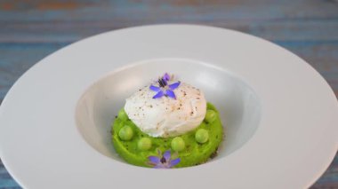 Taze bezelye püresi güzel bir şekilde kaplanmış, ortasında haşlanmış bir yumurta ve borage çiçekleriyle süslenmiş. Yemek canlı renkler ve dokular sergiliyor, her bahar menüsü için mükemmel..