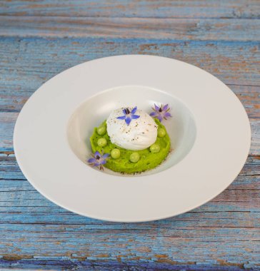 Taze bezelye püresi güzel bir şekilde kaplanmış, ortasında haşlanmış bir yumurta ve borage çiçekleriyle süslenmiş. Yemek canlı renkler ve dokular sergiliyor, her bahar menüsü için mükemmel..