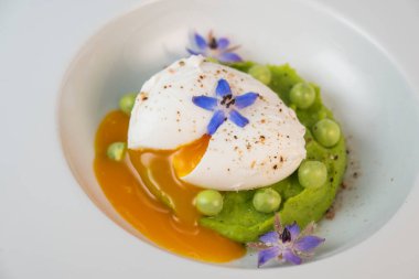 Taze bezelye püresi güzel bir şekilde kaplanmış, ortasında haşlanmış bir yumurta ve borage çiçekleriyle süslenmiş. Yemek canlı renkler ve dokular sergiliyor, her bahar menüsü için mükemmel..