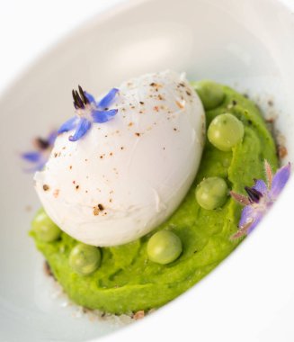 Taze bezelye püresi güzel bir şekilde kaplanmış, ortasında haşlanmış bir yumurta ve borage çiçekleriyle süslenmiş. Yemek canlı renkler ve dokular sergiliyor, her bahar menüsü için mükemmel..
