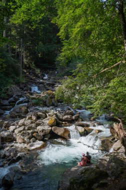 Bir çift, Pont dEspagne yakınlarında, dağlanmış kaya ve yeşilliklerle çevrili, çağlayan bir derenin kenarında oturarak, doğanın güzelliğini yansıtan huzurlu bir anın tadını çıkarıyor..