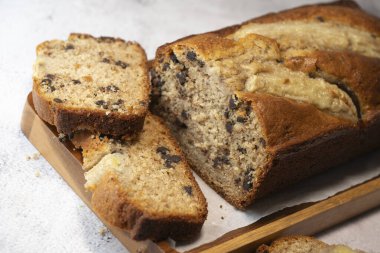 Freshly baked banana bread is sliced and displayed on a wooden cutting board. The bread has a golden-brown crust and chocolate chips, perfect for a cozy snack at home.