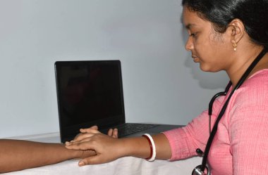 Genç Hintli kadın hemşire beyaz arka planda izole edilmiş parmaklarıyla yaşlı adamın nabzını ölçüyor. Masada tıbbi kontrol, seçici odaklanma.