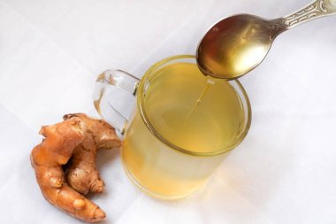 Birisi gümüş bir kaşık tutuyor ve beyaz arka planda zencefil, limon ve zerdeçal ile yapılmış bitki çayıyla dolu bir bardağa bal döküyor. Kopyalama alanı ile seçmeli odak.