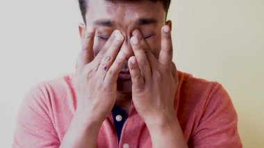 Genç Hintli bir adam tişört giyiyor. Yorgunluk ve baş ağrısı için gözlerini ovuyor. Beyaz arka planda uykulu ve yorgun bir ifade var. Görme sorunu. Seçici odak.