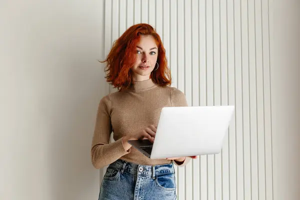 stock image A redhead girl with curly hair poses with a laptop, set against a minimalistic, light wall