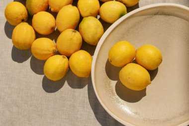 Bir tabakta duran üç taze limonla bir yüzeye dağılmış limonlar, parlak sarı derilerini sergiliyorlar.