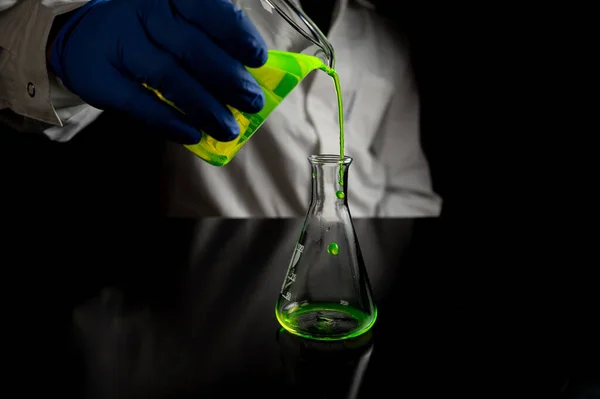 A research scientist experimenting with a green fluorescent droplets in a glass conical flask in dark biomedical laboratory for health care medicine development. Copy space black background