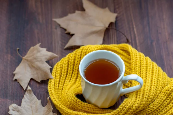 黄色のウールのスカーフと木のテーブルの上の紅葉に囲まれた熱いお茶とカップ — ストック写真