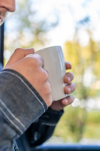 man\'s hand holding a cup of coffee next to a window with autumn background outside