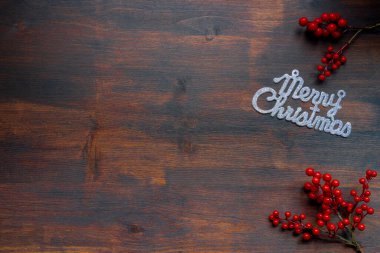 Üzerinde gümüşten parıltılı mutlu noeller yazan bir poster. Koyu ahşap bir arka planda, kırmızı böğürtlen dallarıyla. 