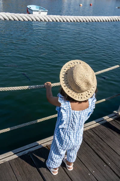 stock image  little girl in a straw hat in the harbour looking at the boats and fish in the sea