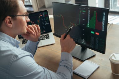 Konsantre iş adamı bilgisayar monitörüne borsa kripto diyagramı ile bakıyor, kalemi ekrana doğrultuyor, çeneye dokunuyor para akışının eğilimini analiz ediyor, yeni bir strateji icat ediyor.