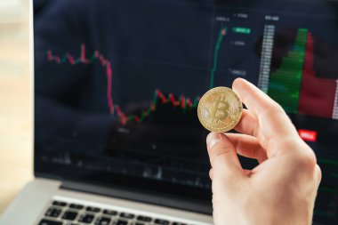 Erkek ellerinin arka planda altın bitcoin tutarken çekilmiş yakın plan resmi. Finansal harita verileriyle birlikte dizüstü bilgisayar ekranı, kripto tüccarı uluslararası değiş tokuşlarda mali veritabanını kontrol ediyor ve kontrol ediyor.
