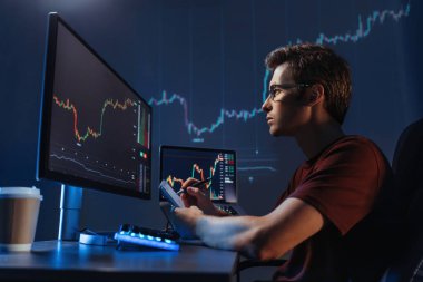Çalışma masasında çalışan genç adam tüccar analistinin yan görünüşü, finansal tabloları, hisse senedi fiyatlarını, hisse senedi fiyatlarını, çevrimiçi ticaret, bilgisayar ekranında kripto para grafiği verilerini kontrol etmek, notlar almak.