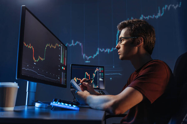 Side view of young man trader analyst at working table, analyzing financial chart, stock quotes, share prices, trading online, checking data on cryptocurrency graph on computer screen, making notes