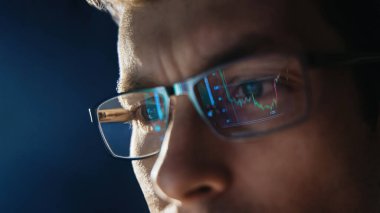 Close up view of focused trader wears eyeglasses looking at pc screen with stock market charts reflection, working online late night. Man analysing crypto market. Selective focus on eye. Copy space. 