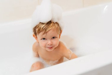 Köpük banyosunda banyo yapan kafasında köpük şapkası olan sevimli küçük bir çocuk. Boşluğu kopyala