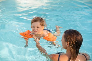 Güneşli bir açık havuzda annesiyle yüzmeyi öğrenirken kollarında yüzen sevimli çocuk gülümsüyor ve yaz eğlencesinin tadını çıkarıyor.. 