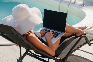 Güneş şapkalı genç bir kadın yüzme havuzunun yanındaki şezlongda dinlenirken dizüstü bilgisayar kullanıyor, dışarıda çalışmaktan zevk alıyor. İş-yaşam dengesi